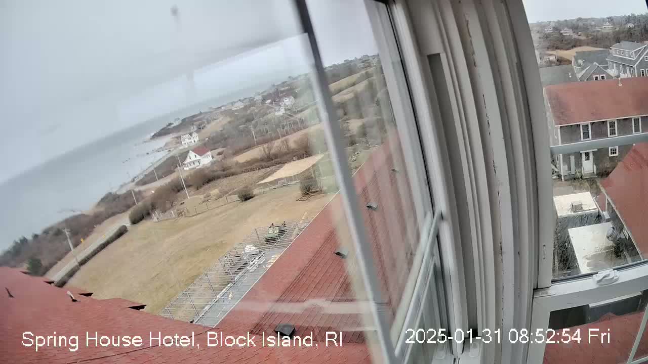 A view from a window shows an overcast day overlooking a coastal town with buildings, open land, and the ocean in the distance.