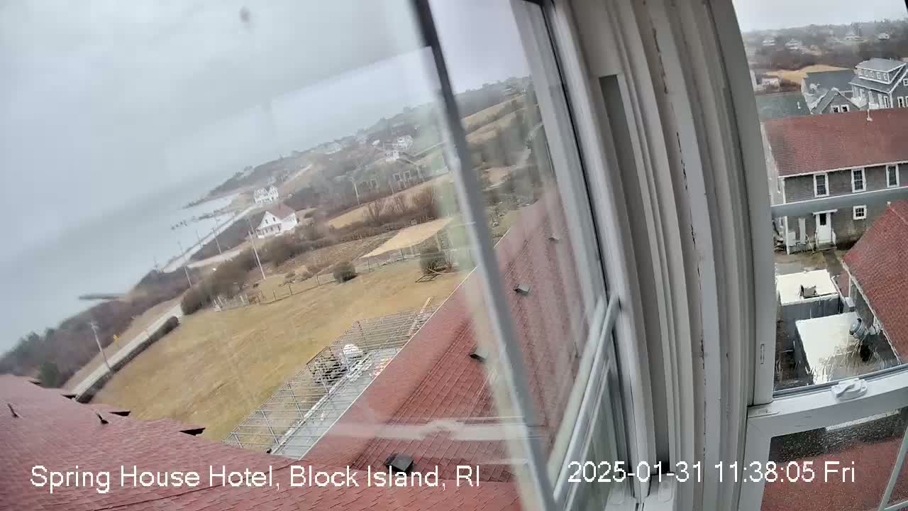 A view from a window shows a cloudy, overcast day overlooking a coastal town with buildings, a road, and open fields.