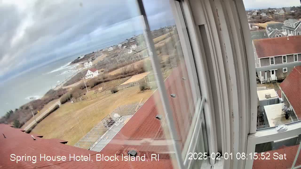 A view from a window shows a cloudy day overlooking a coastal town with buildings, a grassy area, and the ocean with waves visible in the distance.