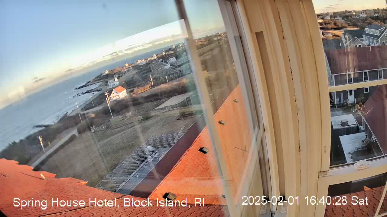 A view from a window shows a calm ocean, a coastal town, and buildings with red roofs under a clear, sunny sky.