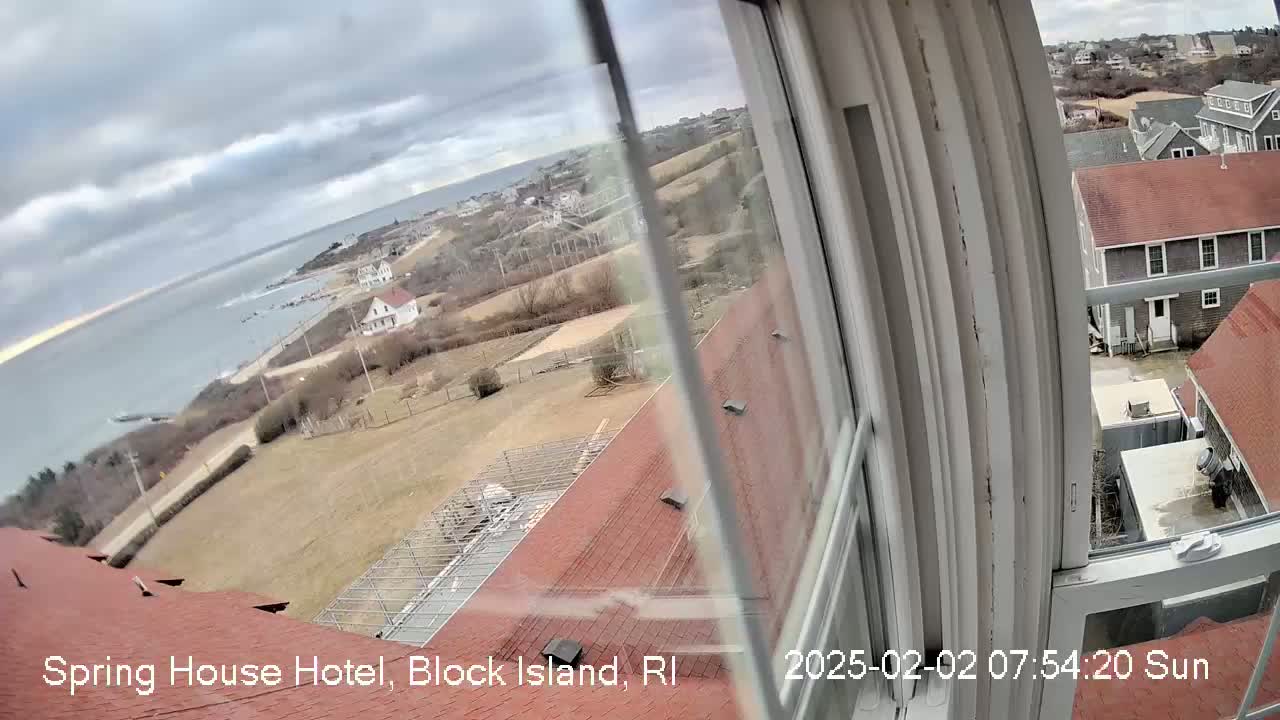 A view from a window shows a cloudy day overlooking a coastal town with red-roofed buildings, a body of water, and brown, sparsely vegetated land.