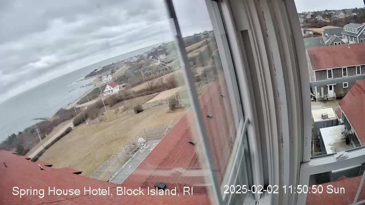 A view from a window shows a cloudy day overlooking a coastal town with buildings, open land, and a body of water under an overcast sky.