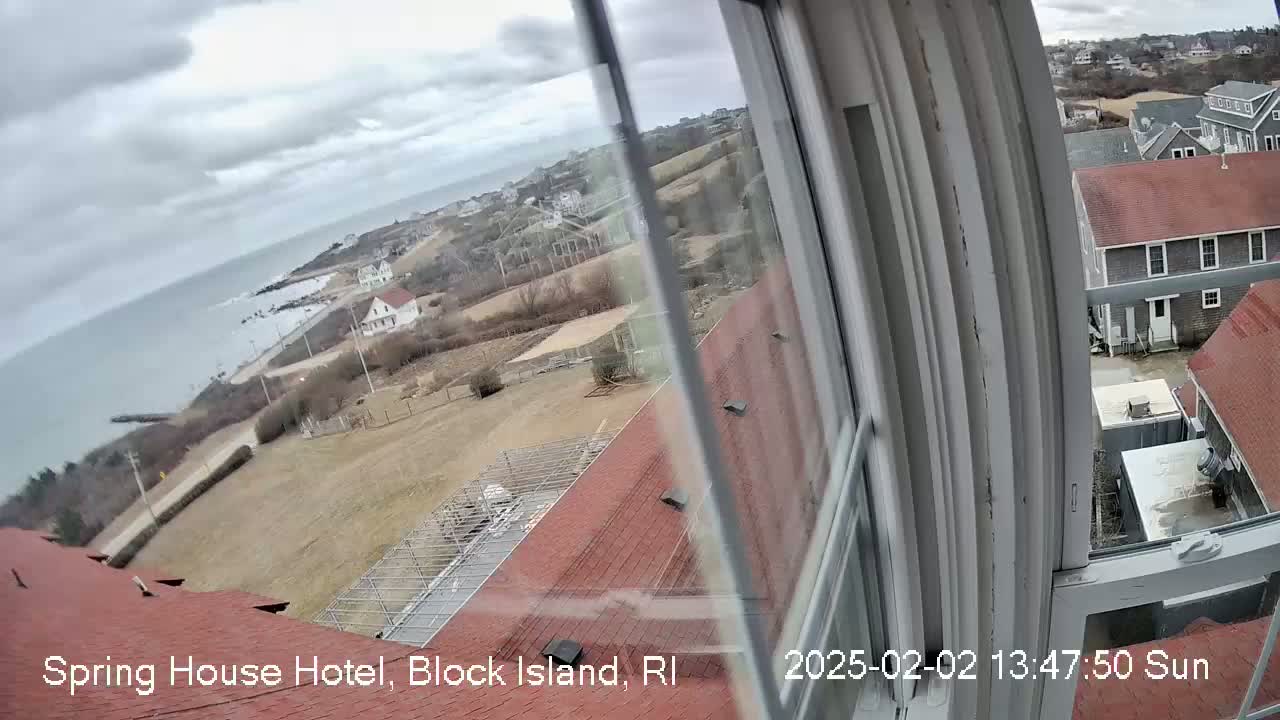 A view from a window shows a cloudy day overlooking a coastal town with red-roofed buildings, brown fields, and a body of water in the distance.