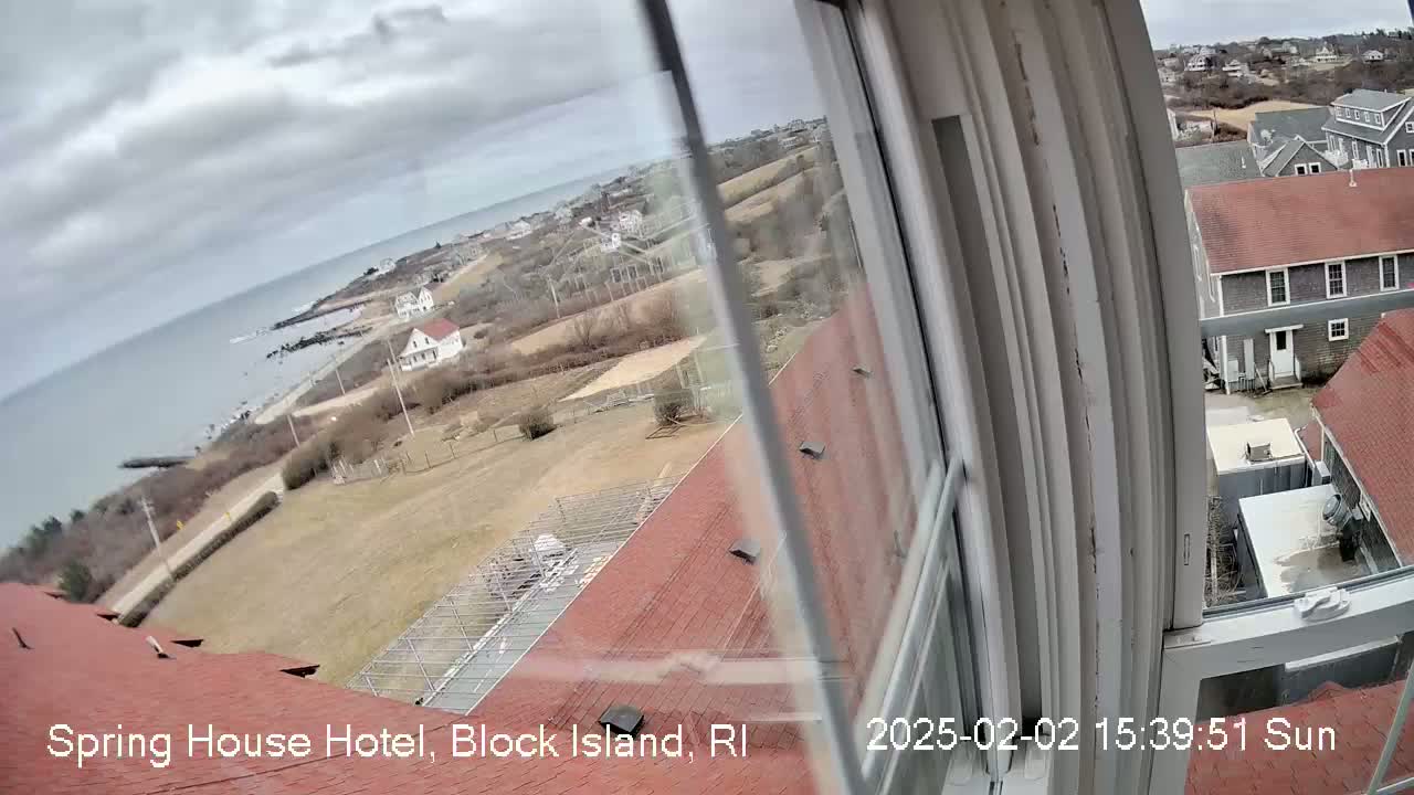 A view from a window shows a coastal scene with overcast skies, a calm ocean, buildings, and brown, grassy land.