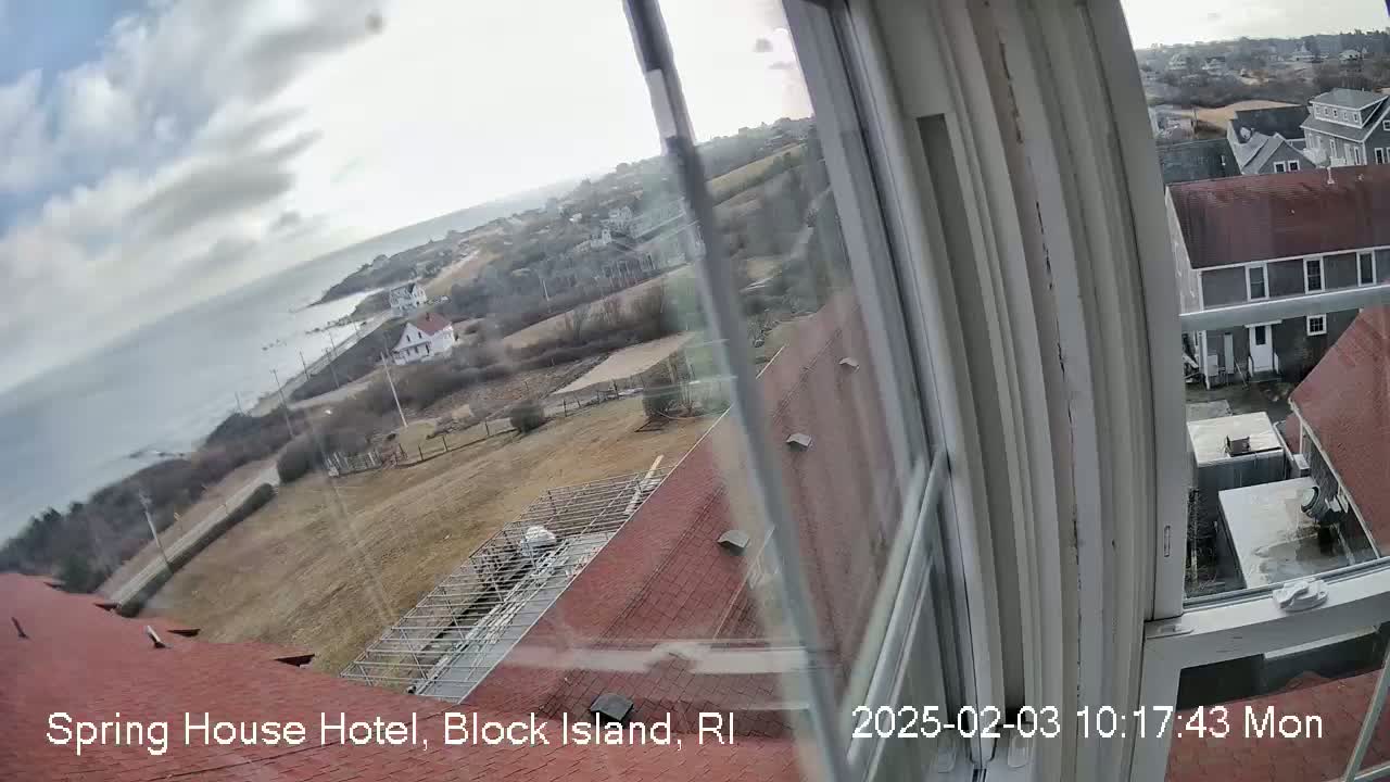 A view from a window shows a partly cloudy day overlooking a coastal town with red-roofed buildings and a body of water in the distance.