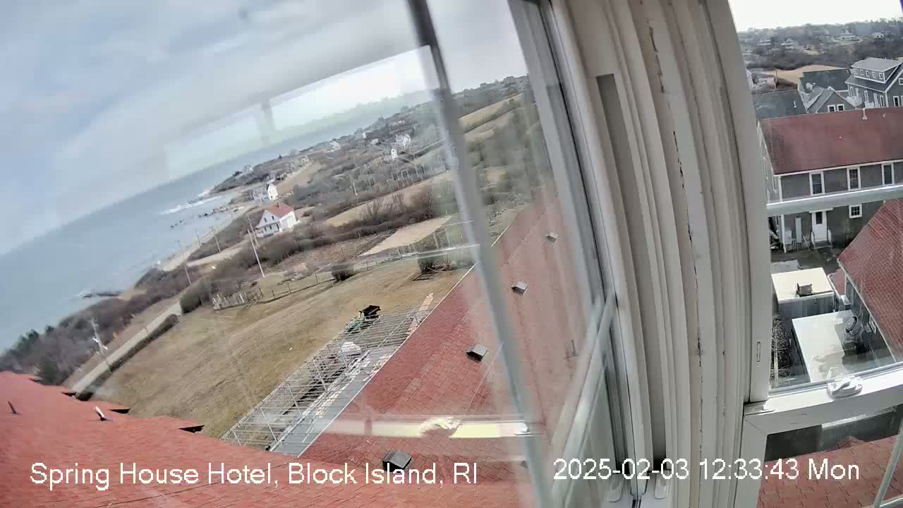 A view from a window shows a coastal scene with overcast skies, a body of water, buildings, and brown grassy areas.