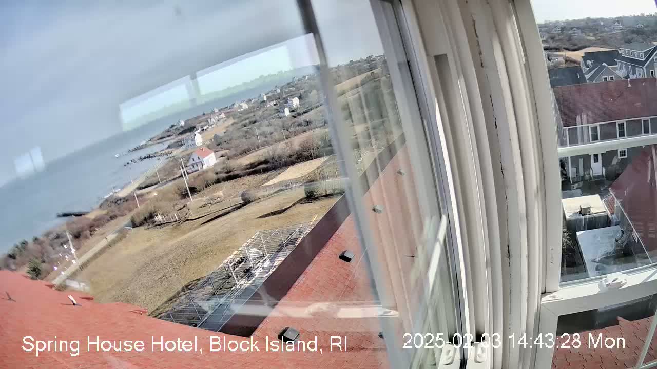 A window view shows a partly cloudy day overlooking a coastal town with houses, a body of water, and some bare, brown land.
