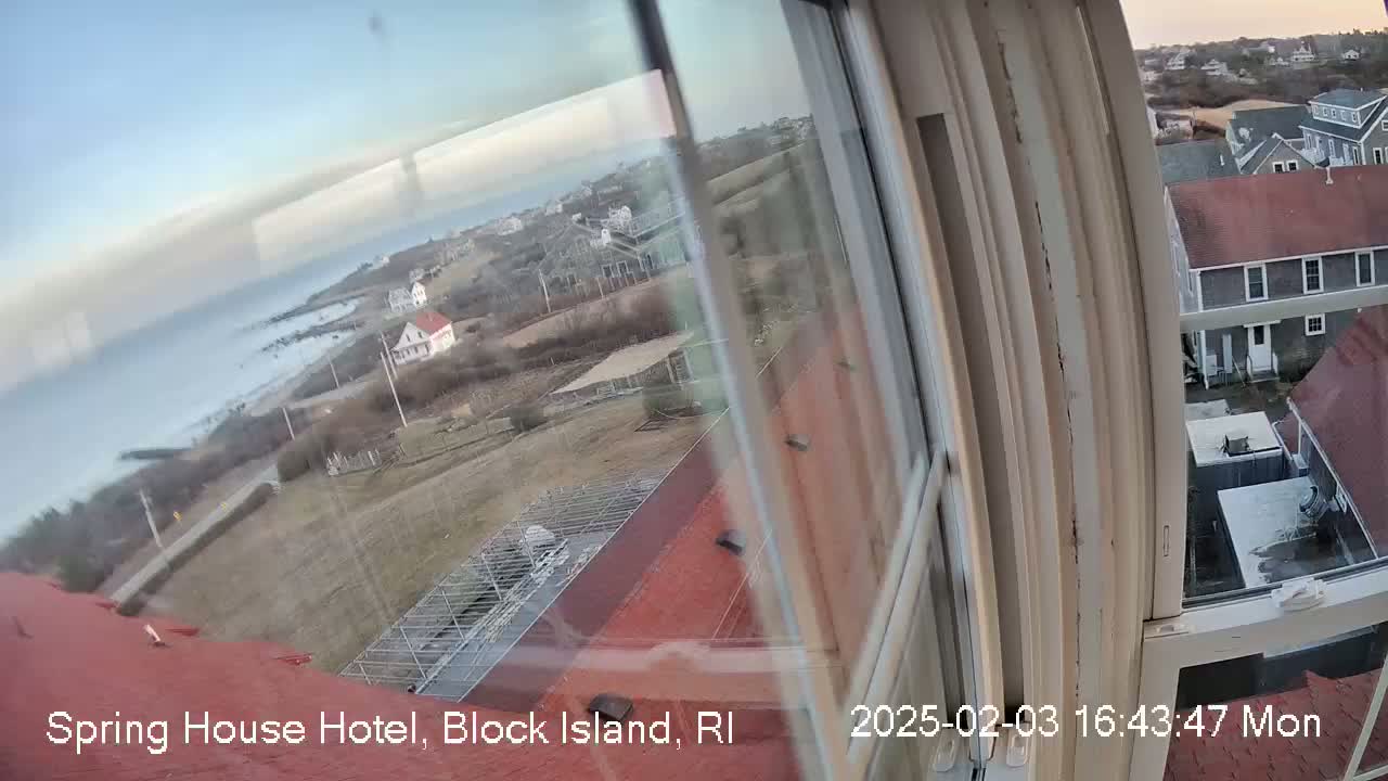 A view from a window shows a partly cloudy sky over a rocky coastline, with buildings and houses situated on a hillside, overlooking the ocean.