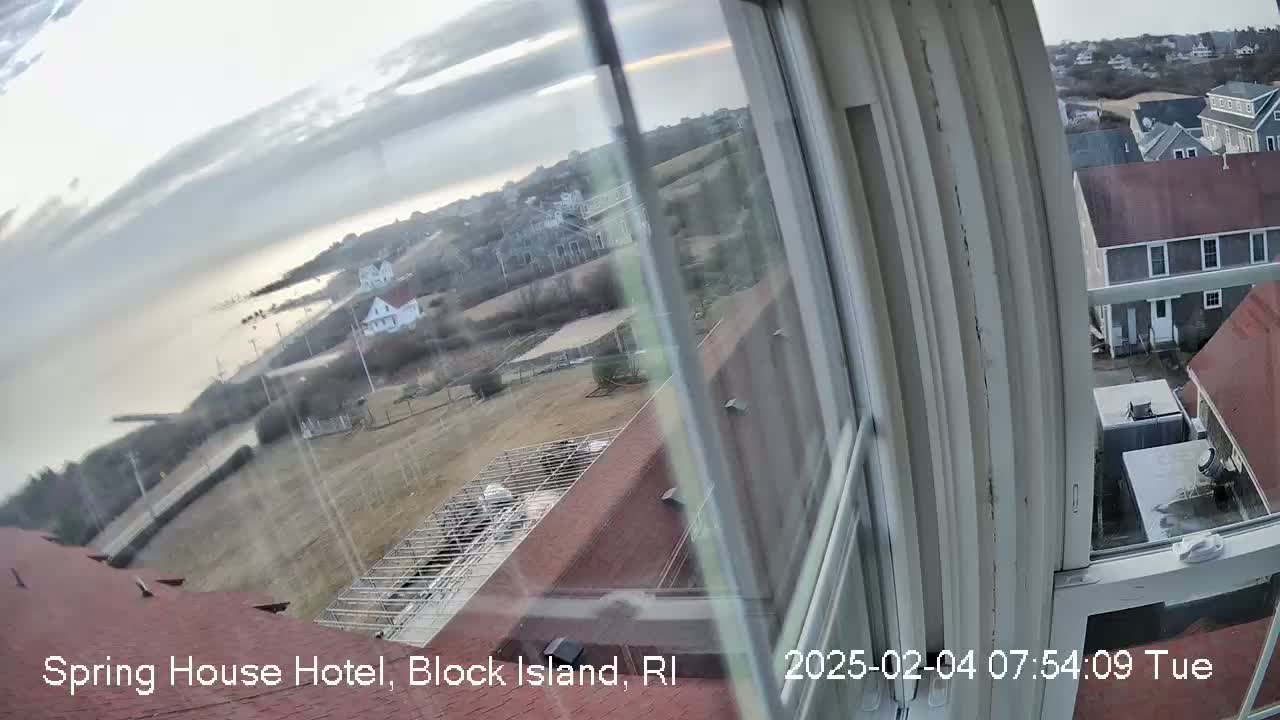 A view from a window shows a calm body of water, houses, and a mostly clear sky with some clouds.