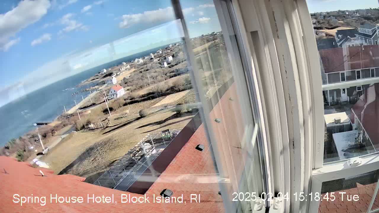 A view from a window shows a partly sunny day overlooking a coastal town with a body of water and a few buildings under a partly cloudy sky.