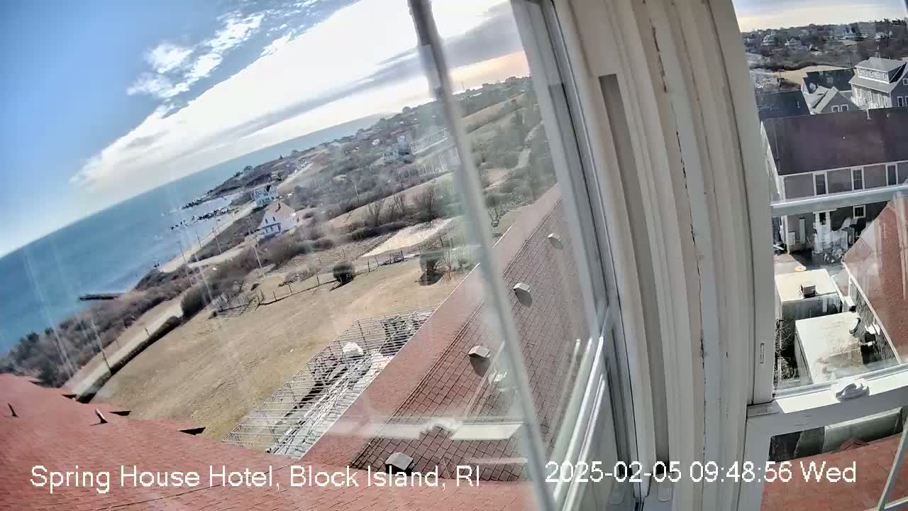 A view from a window shows a mostly sunny coastal scene with a building's red roof in the foreground, overlooking a seaside town and ocean under a partly cloudy sky.