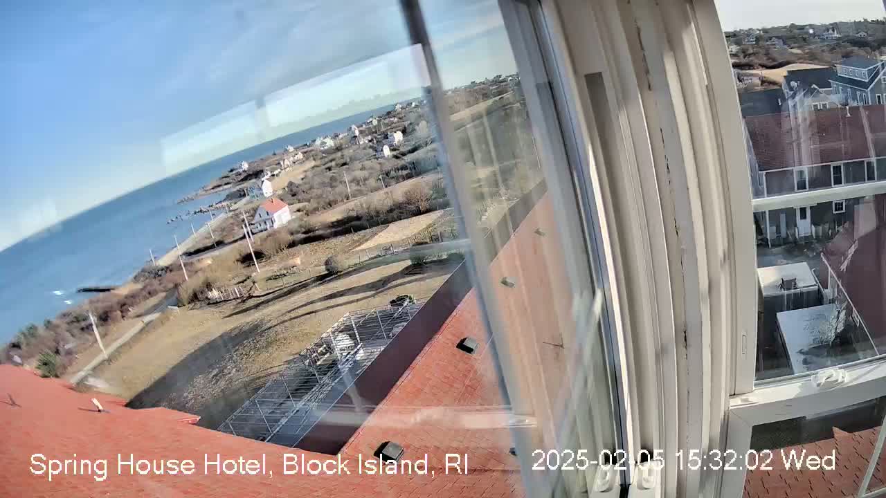A view from a window shows a clear, sunny day overlooking a coastal town with houses, open land, and the ocean.