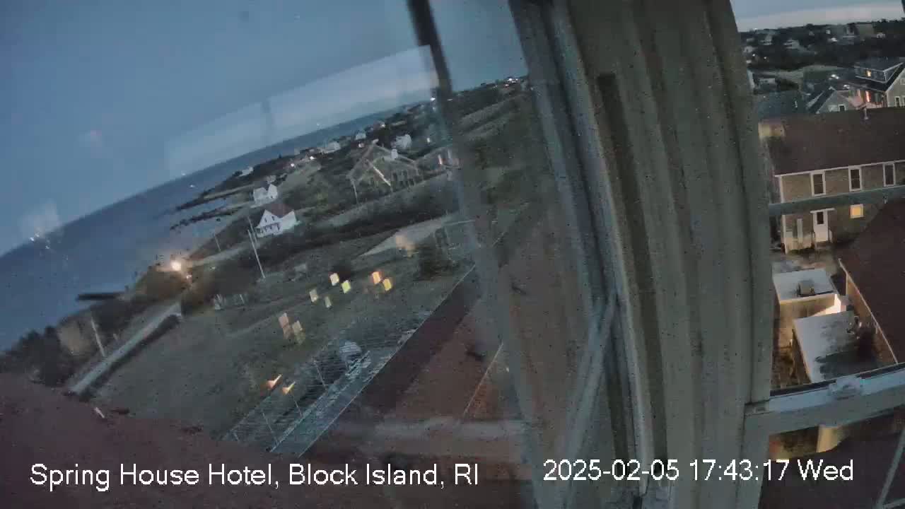 A twilight view of a coastal town, seen from a window, shows a dark ocean and a collection of buildings lit by interior lights.