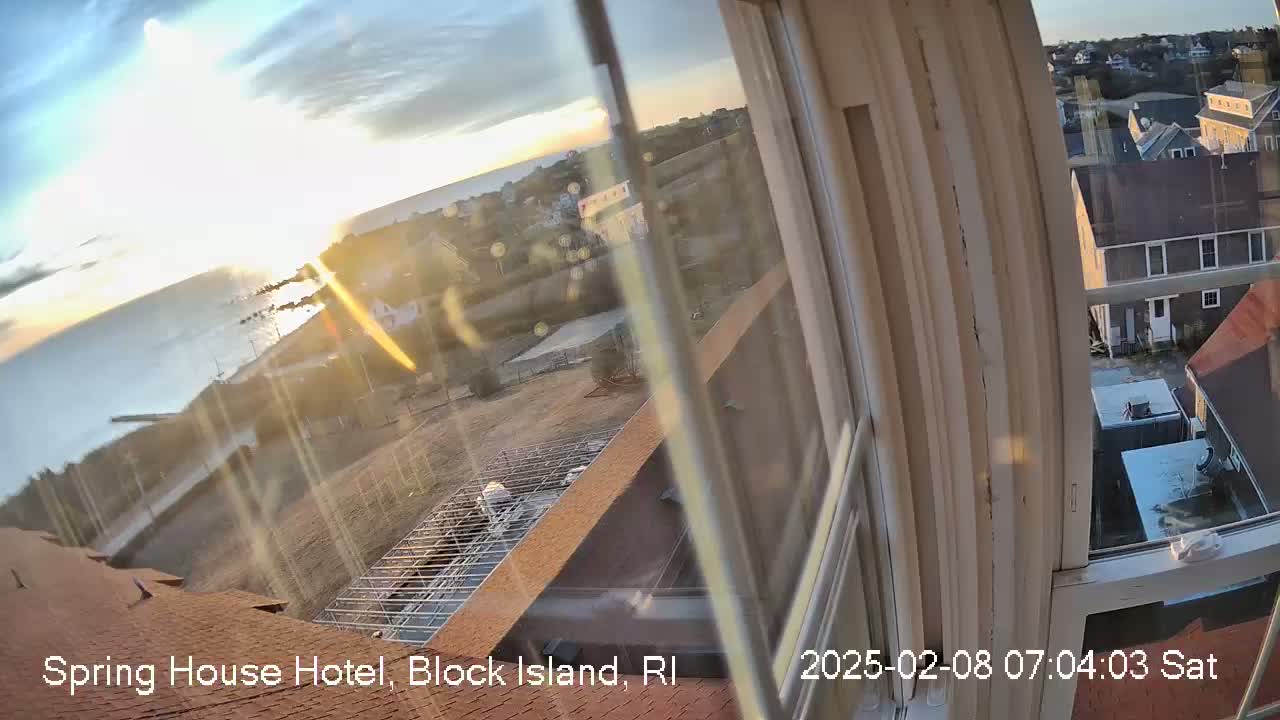 A sunrise over a calm ocean is viewed from a window, showing a partially constructed rooftop and distant houses.