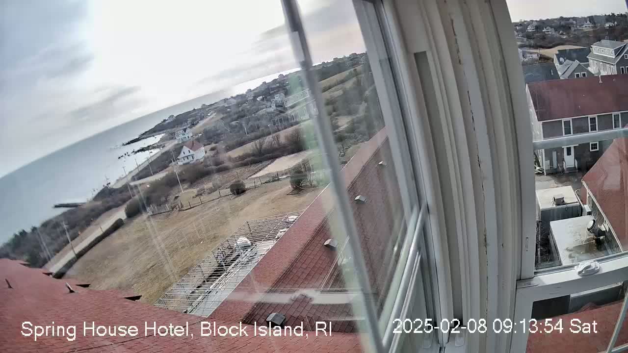 A view from a window shows a cloudy day overlooking a coastal town with brown rooftops, a gray ocean, and barren, brown-green land.