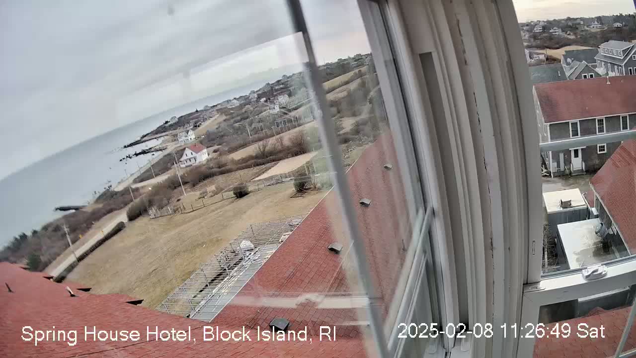 A view from a window shows a coastal town on an overcast day, with a grey ocean in the background and red-roofed buildings in the foreground.