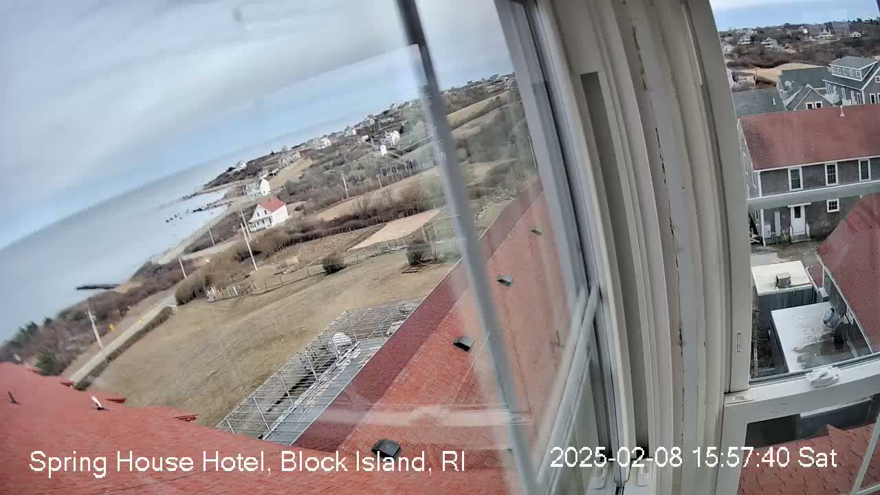 A view from a window shows a coastal scene with overcast skies, featuring a red-roofed building, other buildings, a body of water, and sparsely vegetated land.