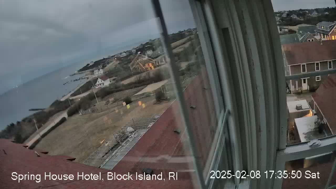 A view from a window shows a coastal town on an overcast day, with buildings, a body of water, and a partially cloudy sky.