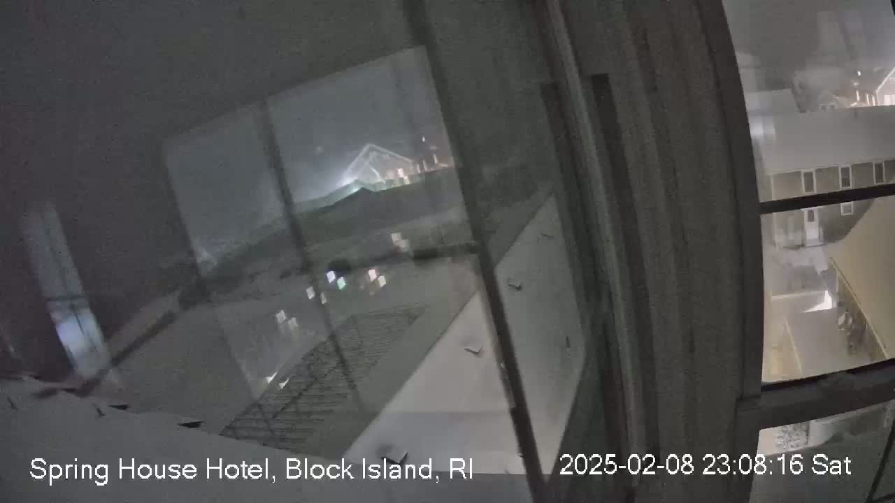 A nighttime view from inside a building shows snow falling outside, with several buildings visible through a window.