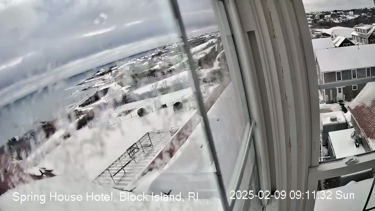A snow-covered landscape viewed from a window, showing a coastal town with houses and a body of water in the distance under a cloudy sky.
