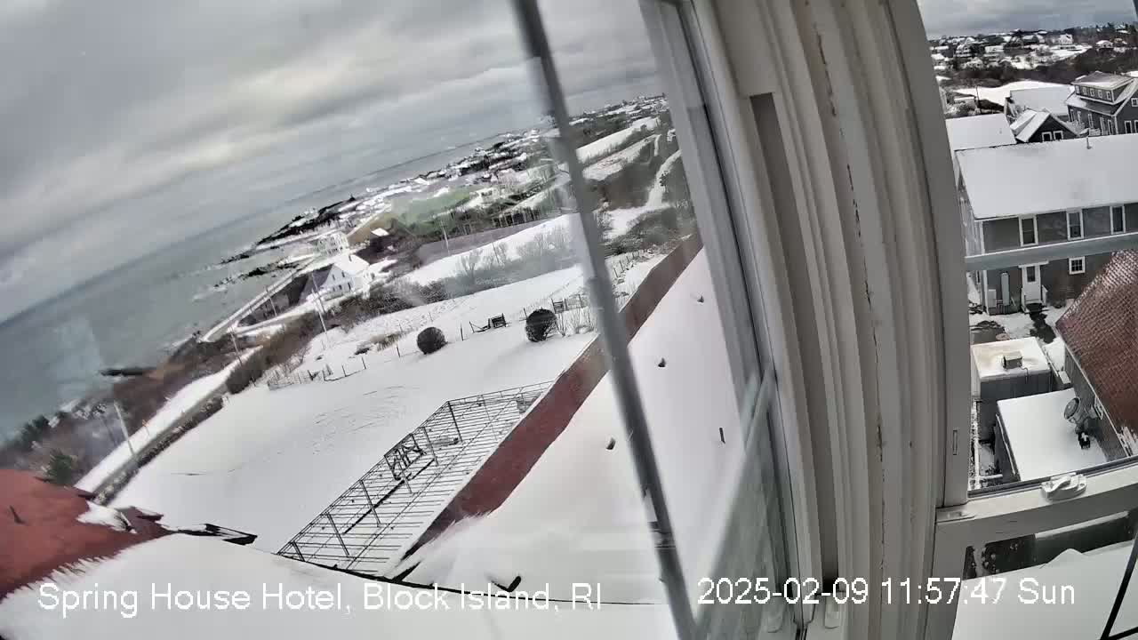 A snowy landscape viewed from a window shows a coastal town with ocean in the distance under an overcast sky.
