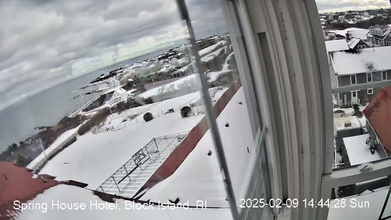 A snow-covered coastal town is seen from a window, with a gray, cloudy sky and a body of water visible in the distance.