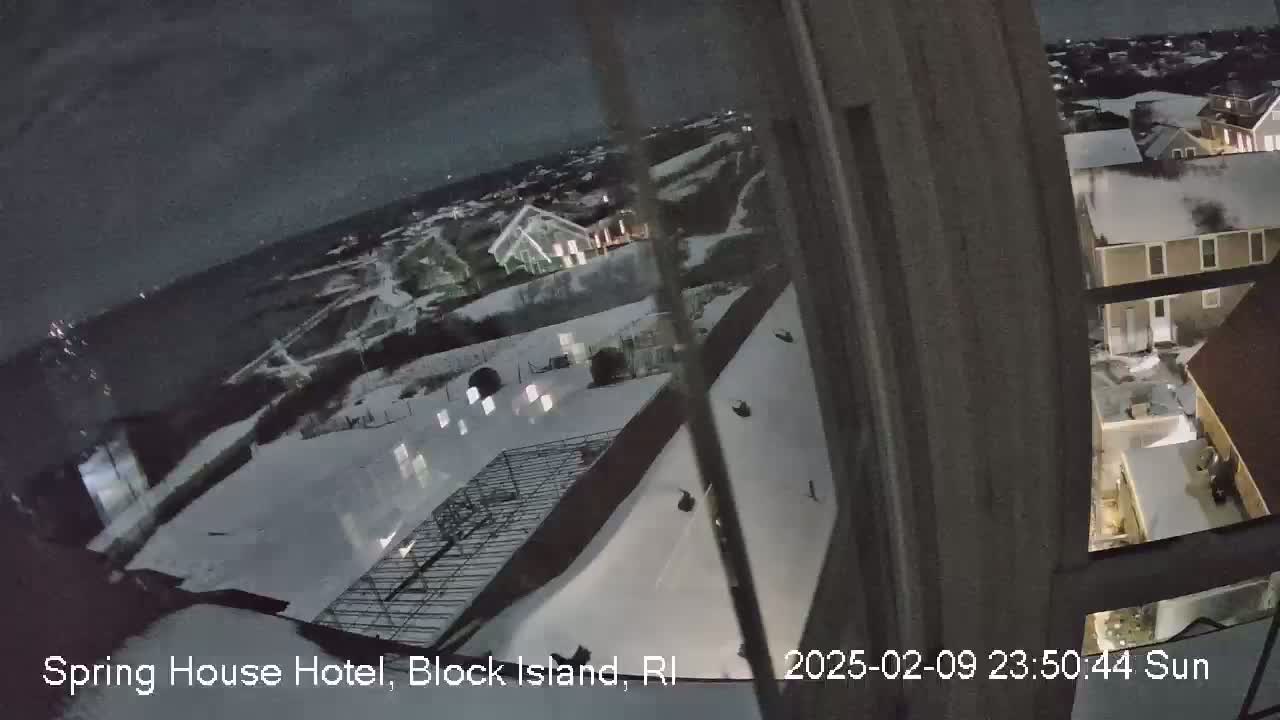 A nighttime view from a window shows a snow-covered landscape with houses and a metal framework structure, under a dark, mostly clear sky.