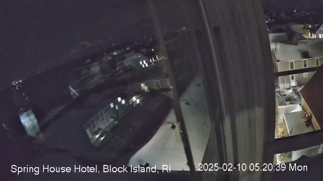A nighttime, overhead view from a window shows a snow-covered residential area with houses and dimly lit streets.