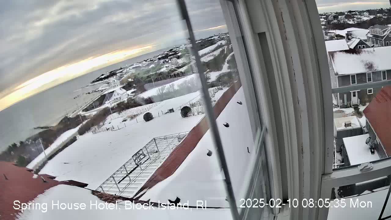 A snow-covered landscape viewed from a window, showing a coastal town and ocean under a partly cloudy sky.