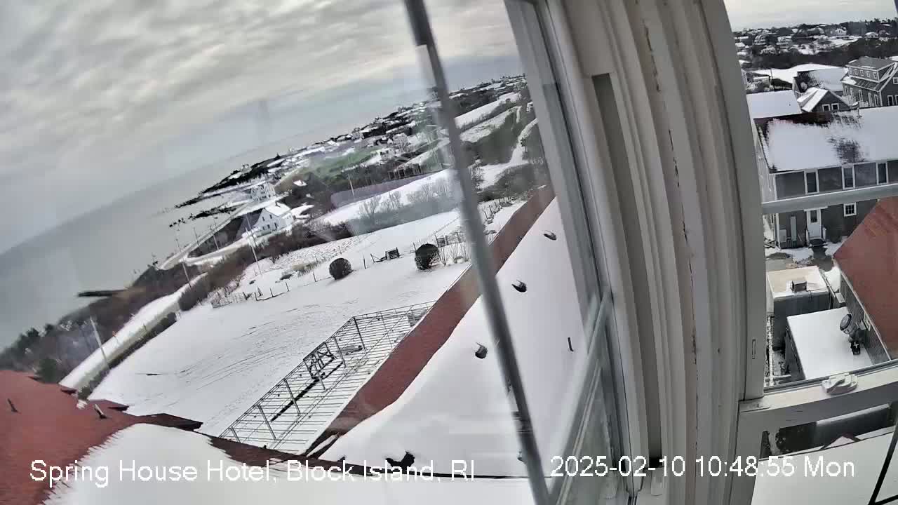 A snow-covered landscape is viewed from a window, showing a coastal town, ocean, and other buildings in the distance under an overcast sky.
