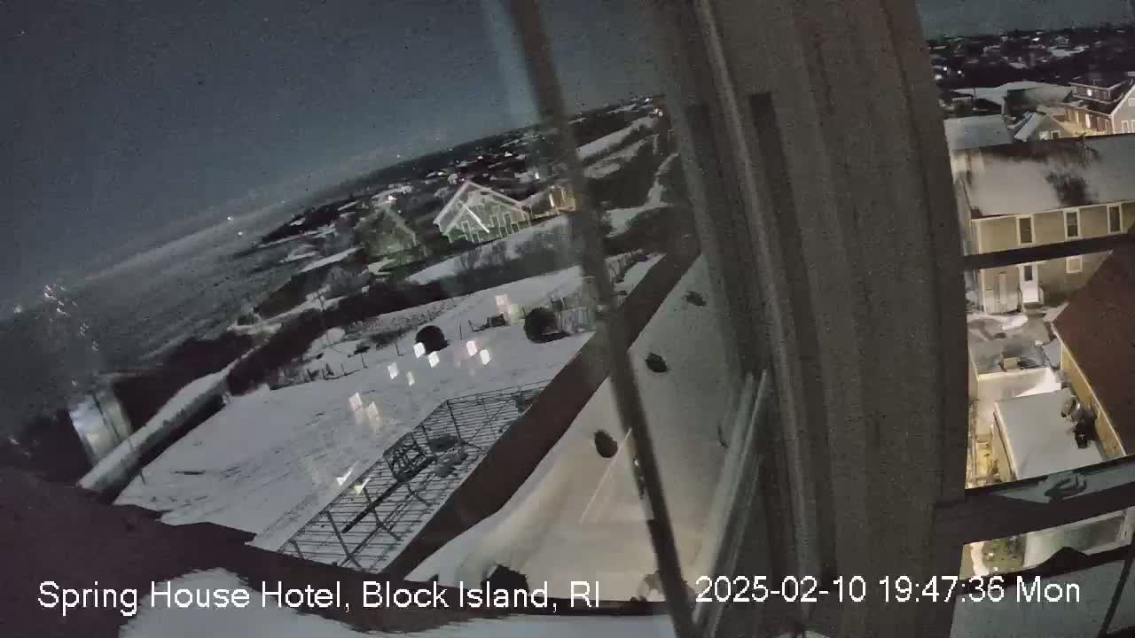 A nighttime view from a window shows a snow-covered landscape with buildings and a dark ocean in the distance under a partly cloudy sky.