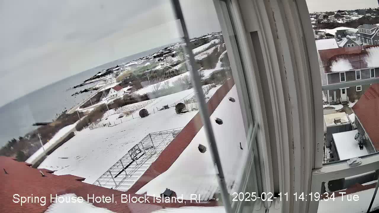 A snow-covered landscape viewed from a window, showing a coastal area with buildings and a body of water in the distance under an overcast sky.