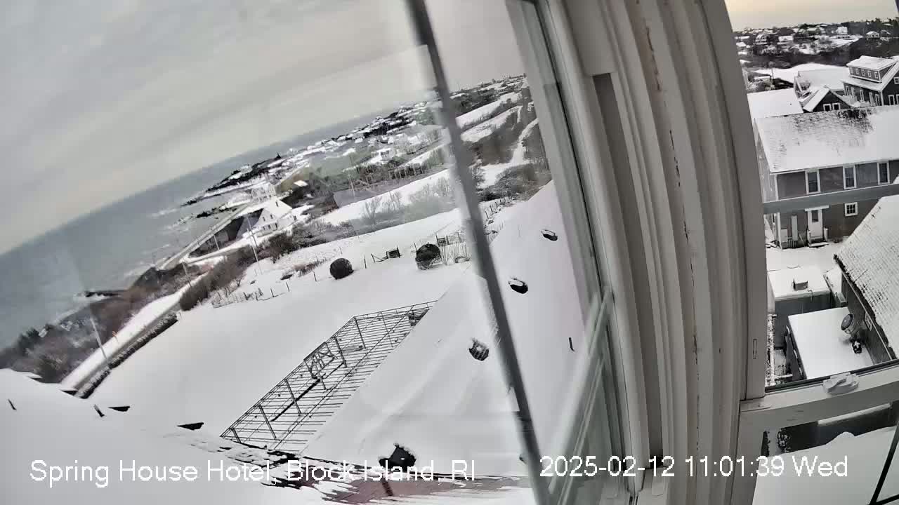 A snow-covered landscape is viewed from a window, showing a coastal scene with houses and a body of water in the distance under a cloudy sky.