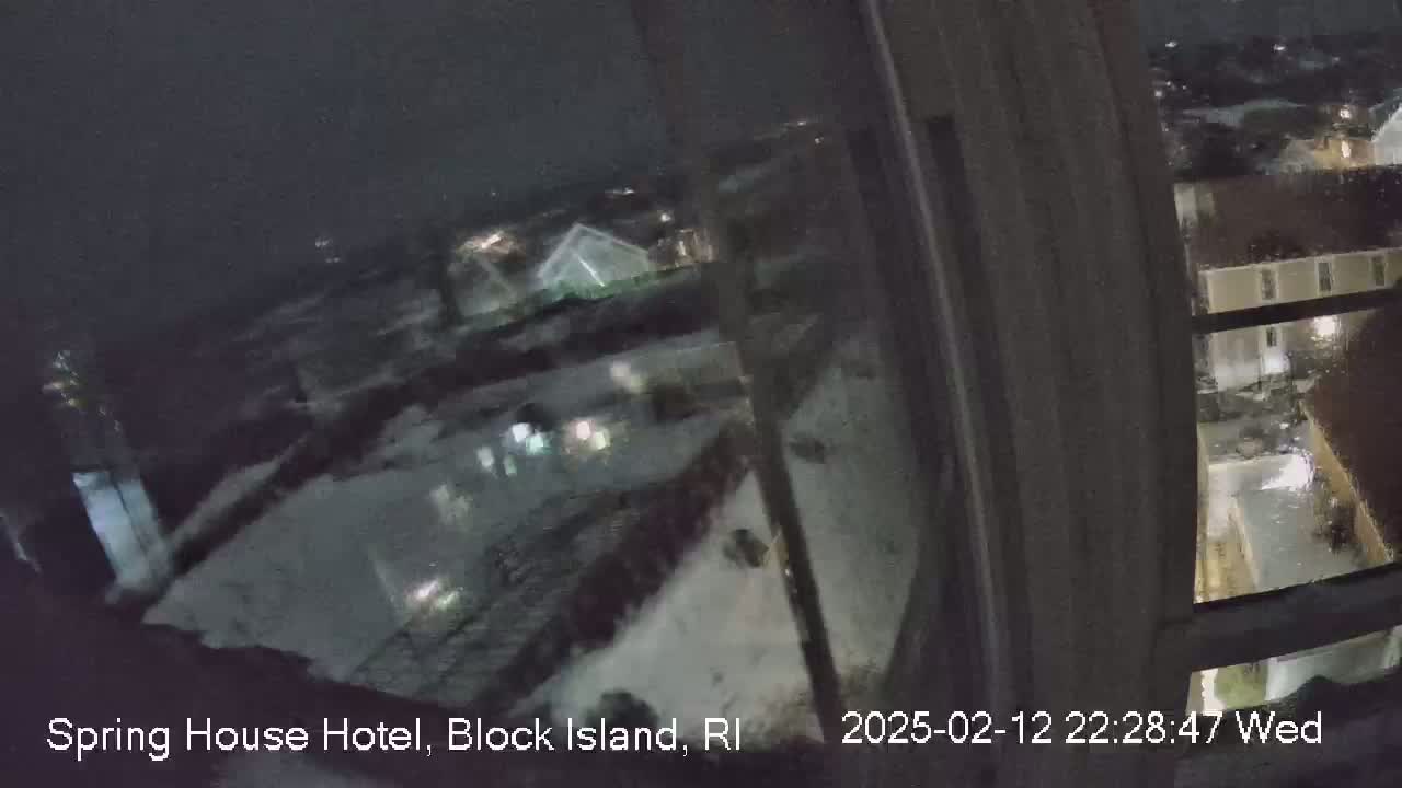 A nighttime view from a window shows snow-covered rooftops and buildings under a dark, snowy sky.