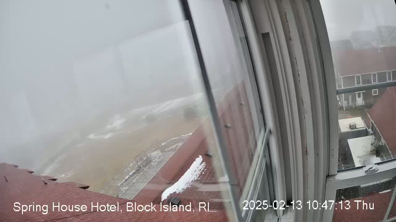 A view from a window shows a foggy, snowy scene of a stormy ocean and nearby buildings.