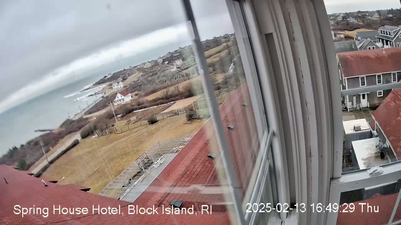 A view from a window shows a cloudy day overlooking a coastal town with red-roofed buildings, a grey ocean, and brown, grassy areas.