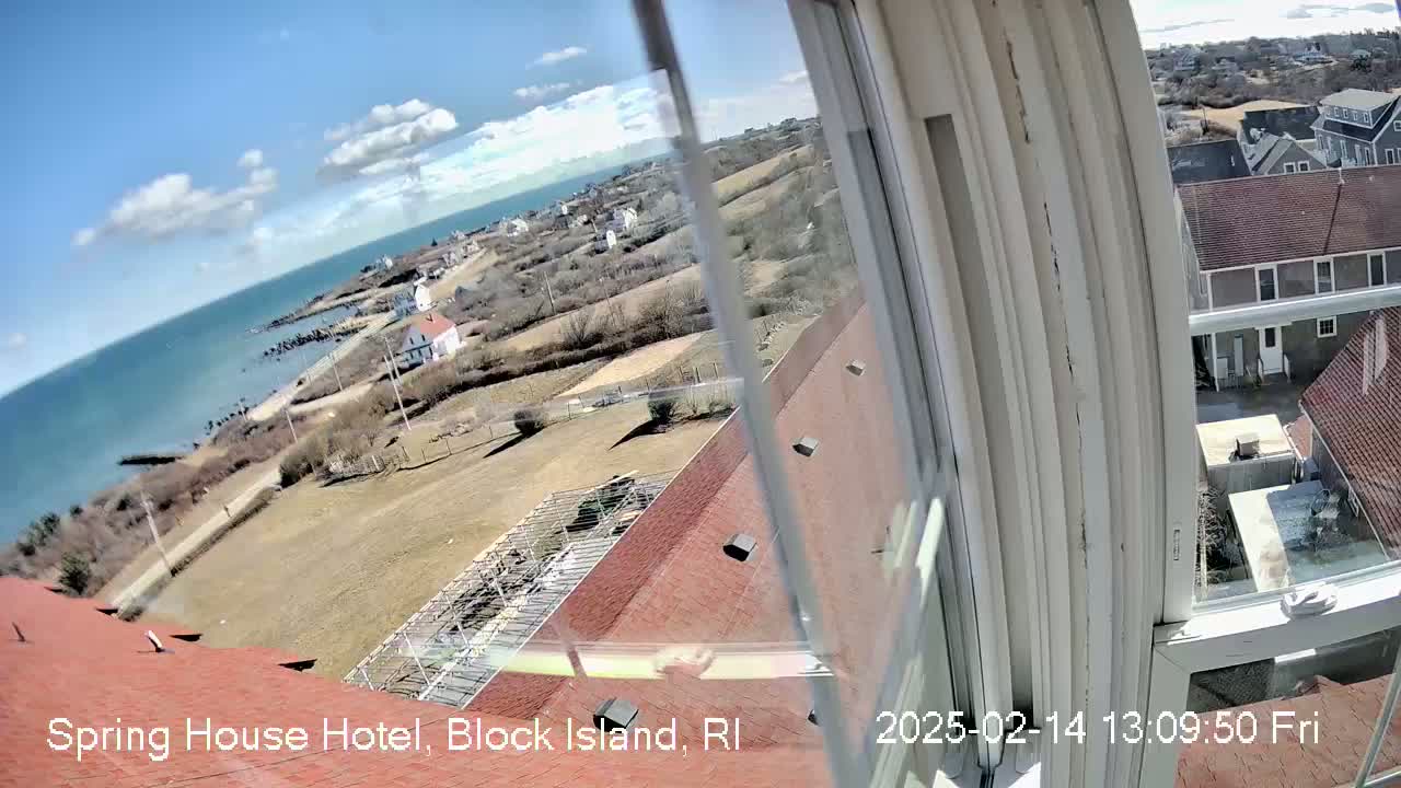 A mostly sunny view from a window shows a coastal landscape with buildings, a rocky shore, and ocean under a partly cloudy sky.