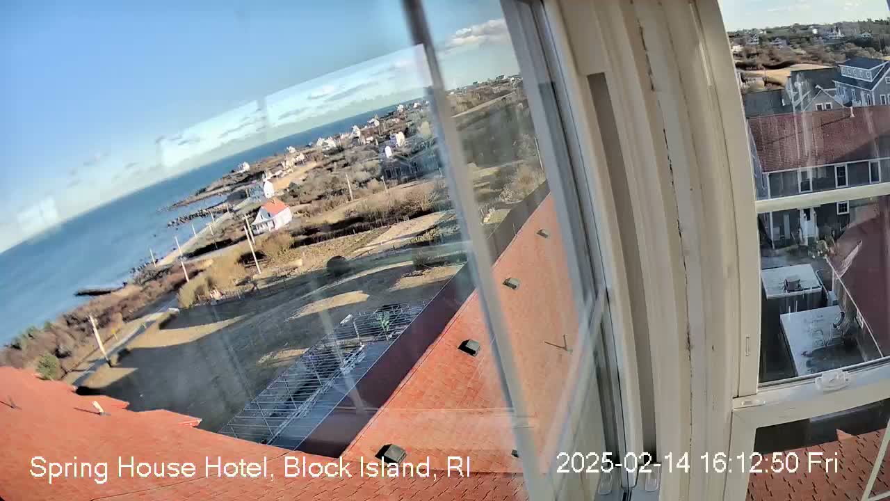 A view through a window shows a sunny coastal town with a rocky shoreline and ocean under a mostly clear sky.