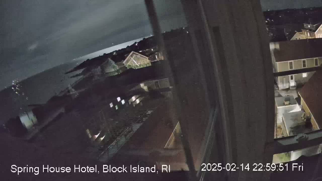 A nighttime view from a window shows a dark body of water, a town with dimly lit buildings, and an overcast sky.
