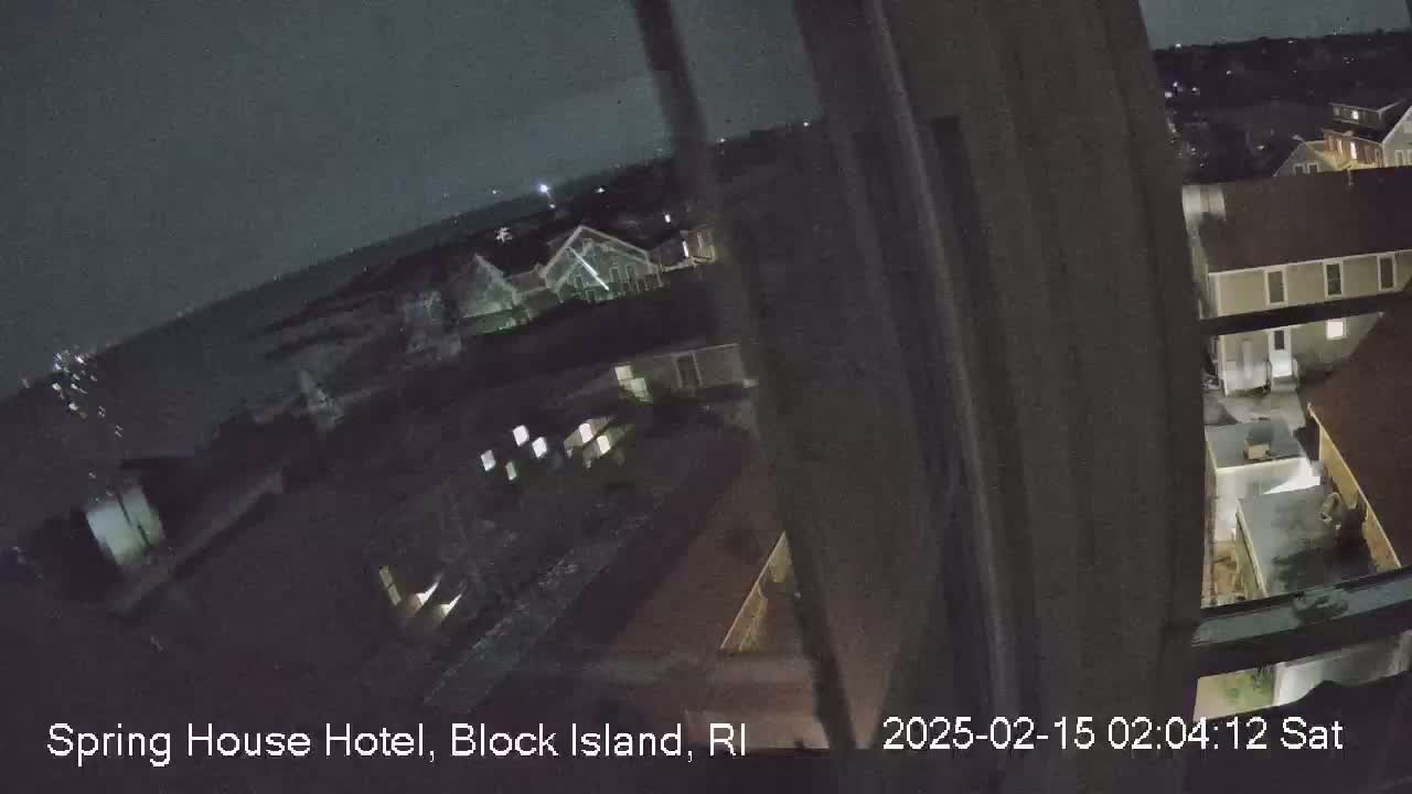 A nighttime, high-angle view from a window shows dark buildings and a body of water in the distance under a cloudy sky.