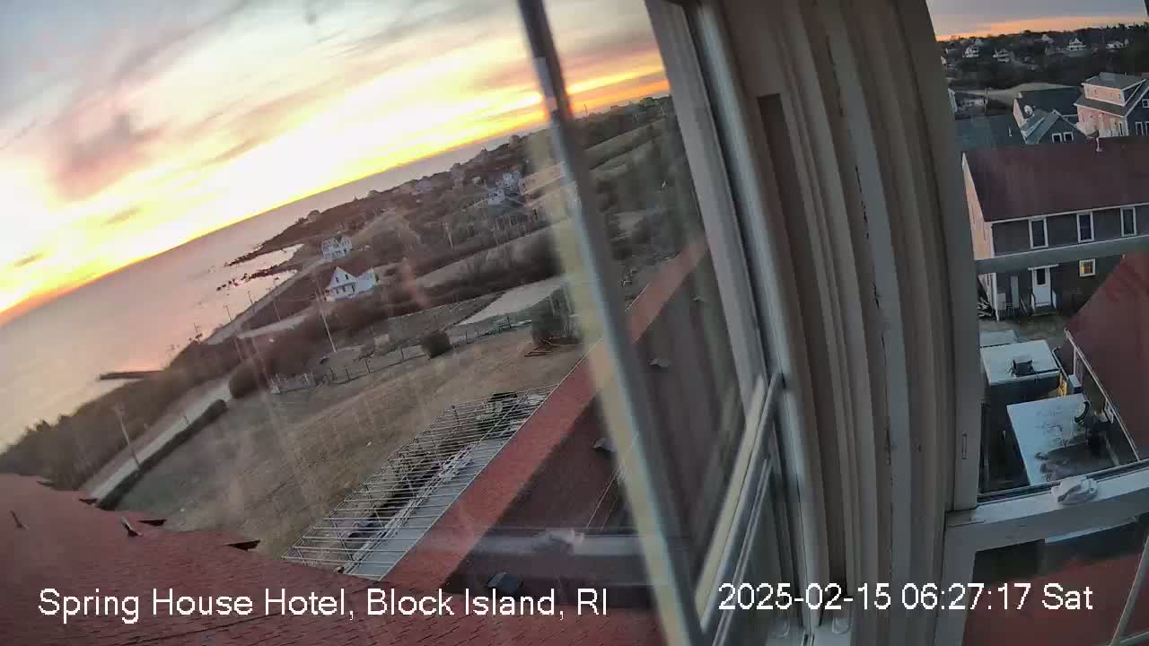 A sunrise over a calm ocean is seen from a window, showing a coastal town with buildings and a partially-constructed rooftop below.