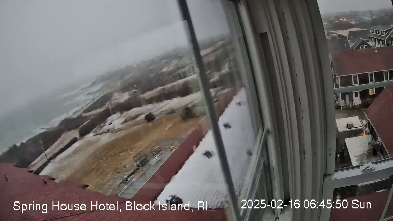 A snow-covered coastal scene is viewed from a window, showing rough ocean waves and buildings in the distance under an overcast sky.