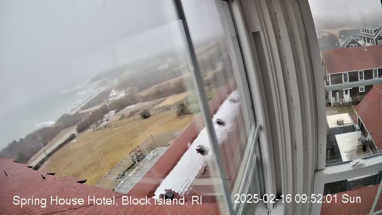A snow-covered rooftop and surrounding landscape are seen through a window, with a blurry, overcast ocean visible in the distance.
