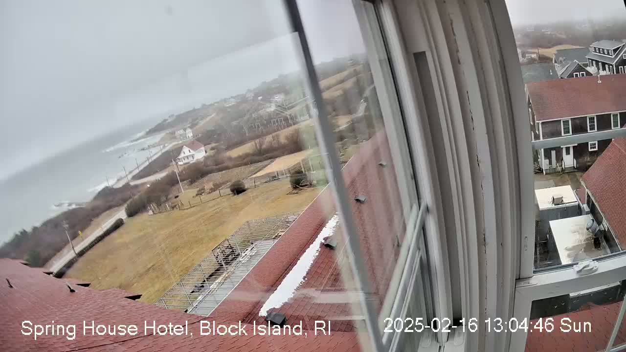 A view from a window shows a coastal town on a cloudy day with some snow on rooftops and a rocky shoreline with a gray ocean visible in the distance.