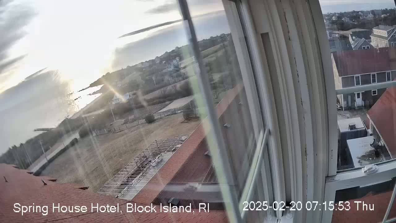 A view from a window shows a partly sunny, possibly cool day overlooking a coastal town with a body of water and buildings, including a partially constructed roof structure.