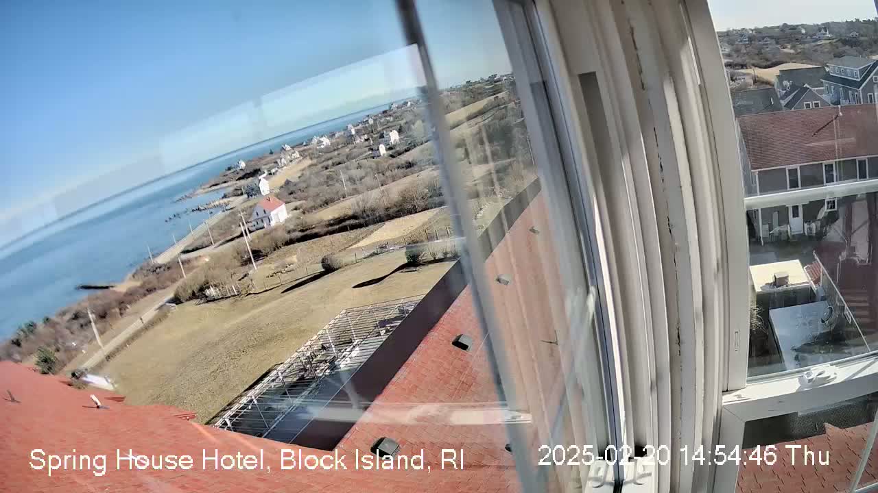 A view from a window shows a sunny coastal scene with a body of water, houses, and brown rooftops under a clear blue sky.