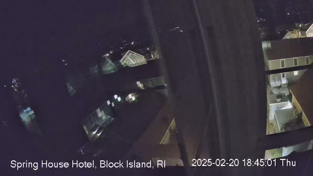 A nighttime, high-angle view from inside a building shows a dark, partially obscured view of several houses and buildings with some lights on.