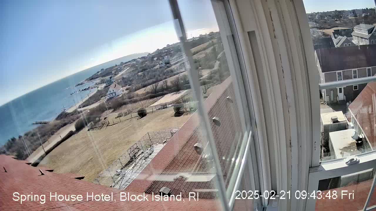 A view from a window shows a coastal town on a sunny day, with ocean visible in the distance and brown, mostly barren land closer to the camera.