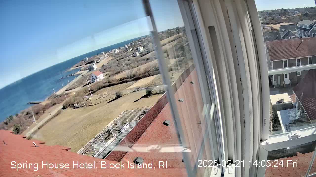A coastal view on a sunny day is seen from a window, showing a seaside town with red-roofed buildings, open land, and a body of water.