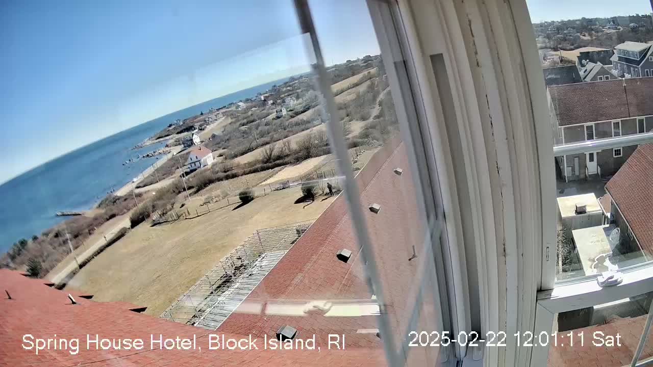A view from a window shows a coastline with a body of water under a clear blue sky, overlooking buildings and a grassy area with some sparse trees.